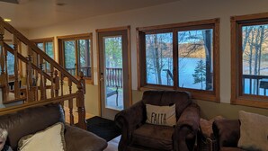 A living room with a brown leather sofa, a brown armchair with a 'FAITH' pillow, a wooden staircase, and large windows with a view of trees and a lake. - Lake Home property (Luck)