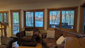 A cozy living room with leather sofas, a wooden coffee table, and large windows with a view of trees and a deck. - Lake Home property (Luck)