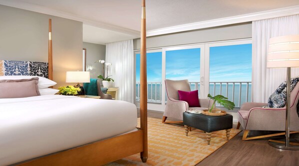 Habitación de hotel con una cama grande, un área de descanso con una silla y una mesita, y vista al mar a través de amplios ventanales.