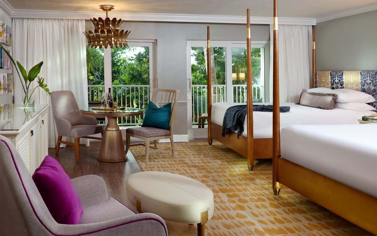 A hotel room with a four-poster bed, a sitting area with a table and chairs, and a balcony with a view of greenery.