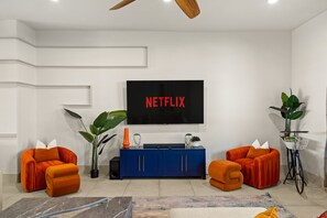 Uma sala de estar moderna com uma televisão de tela plana exibindo 'NETFLIX' em letras vermelhas, duas poltronas laranjas, um armário azul e um vaso de plantas.