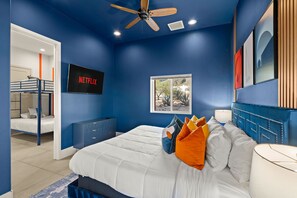 Une chambre avec un lit superposé, un lit avec des oreillers orange, un ventilateur de plafond et un téléviseur affichant le logo « NETFLIX ».