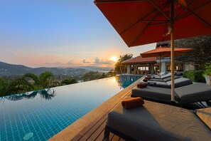 An infinity pool with lounge chairs and umbrellas, overlooking a scenic landscape with mountains and a sunset. - Samui Summit Villa : 4 Bedroom, Panoramic Bliss (Chang Wat Surat Thani)