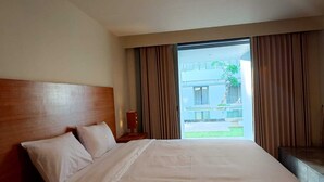 A hotel room with a large bed, a wooden headboard, and a view of a building and greenery outside. - Kuta Living (Kuta)