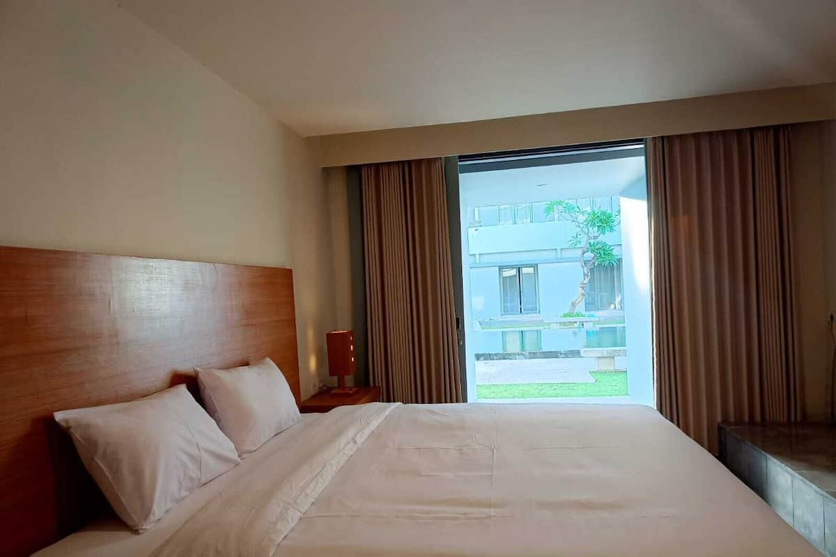 A hotel room with a large bed, a wooden headboard, and a view of a building and greenery outside.