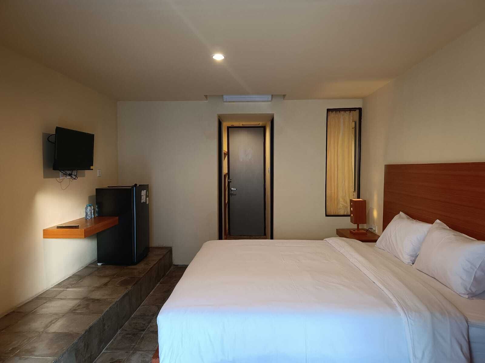 A hotel room with a bed, a television mounted on the wall, a small refrigerator, and a door with a curtain.
