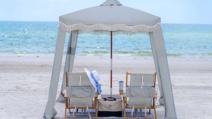 Un cabanon de plage avec un tissu à rayures et des franges, deux chaises en bois, une table avec un chapeau et un sac, et une vue sur la mer.