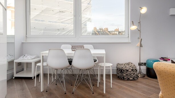 Een moderne kamer met een witte tafel en stoelen, een groot raam met uitzicht op gebouwen, een vloerlamp en een gestreept kruikzak.
