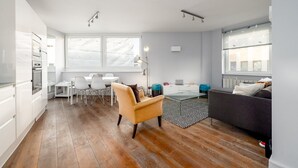 Een moderne keuken met witte kasten, een eethoek met een witte tafel en stoelen, een kamerkleed met een patroon en een grijze bank.