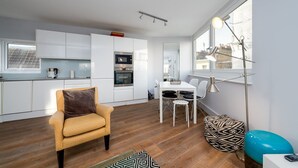 Een kleine keuken met witte kastjes, een magnetron en een koffieapparaat. Er is een eethoek met een witte tafel en stoelen, en een bank met een decoratief kussen.