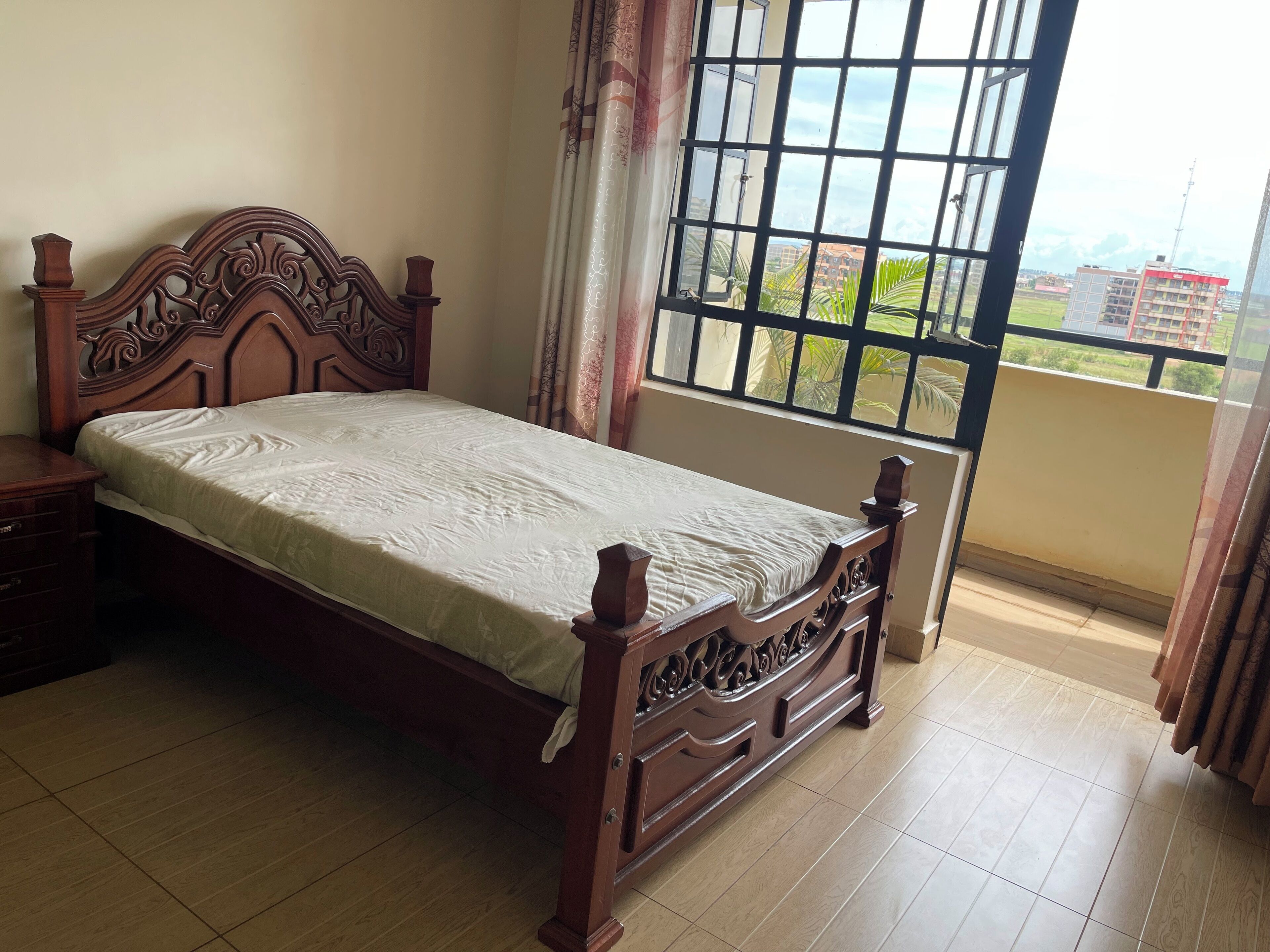 A bedroom with a wooden bed, a nightstand, a window with a view of buildings, and a curtain.