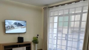 Een woonkamer met een flatscreen televisie, een houten tv-meubel, beige banken, een wit vloerkleed en een groot raam met dunne gordijnen.