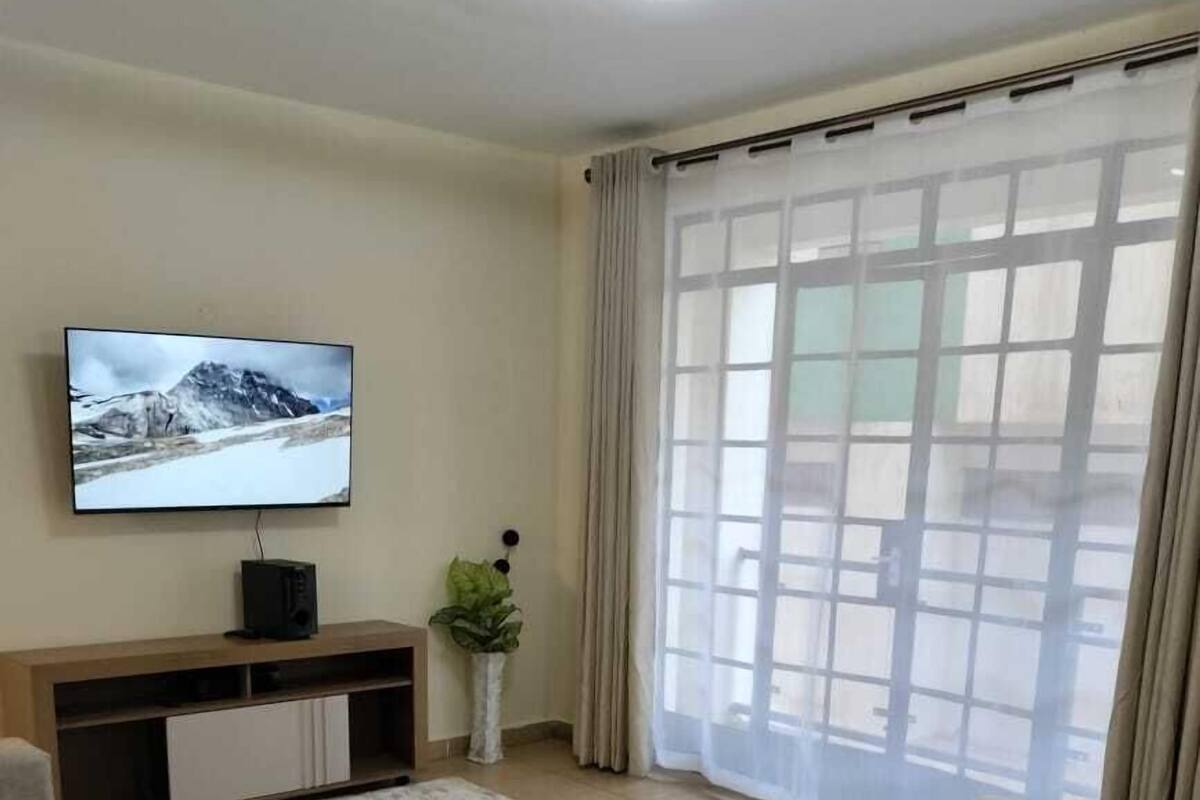 Una sala de estar con un televisor de pantalla plana, un mueble de madera para equipo de entretenimiento, sofás color beige, un tapete blanco y un ventanal con cortinas de gasa.