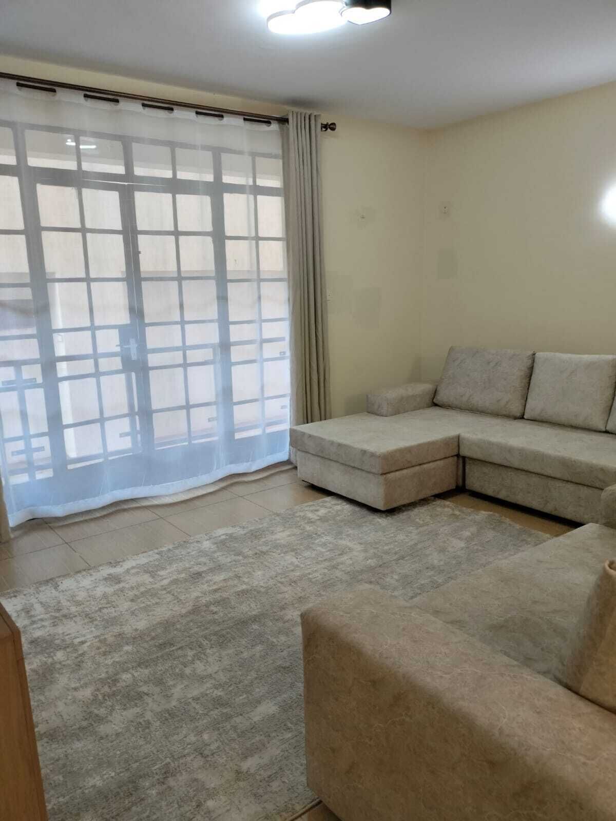 Un salon avec un canapé d’angle beige, une grande fenêtre ornée d’un rideau léger et un sol carrelé.