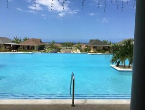 Ein Pool in einer Ferienanlage mit klarem, blauem Wasser, umgeben von Palmen und Häusern mit geneigten Dächern.