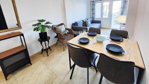 Comedor con mesa de madera dispuesta para cuatro personas, rodeada de sillas oscuras. También se ve una silla de mimbre y una mesita con una planta en maceta.