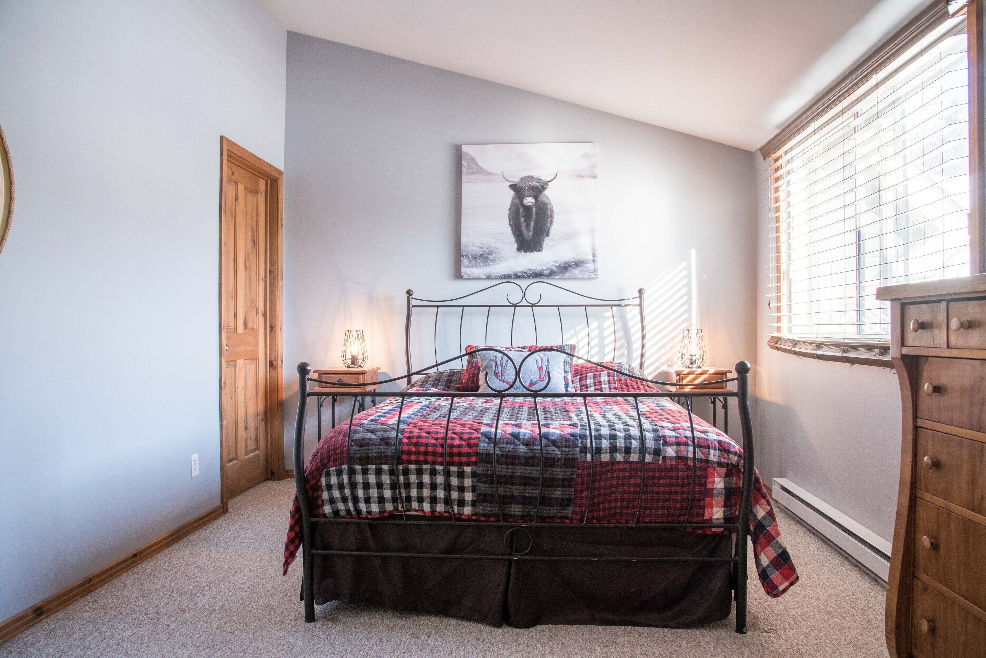 A bedroom with a bed, a wooden door, a window with blinds, a wall-mounted picture, and a dresser.