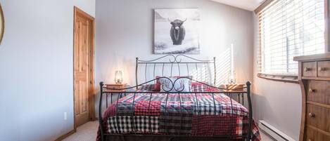 A bedroom with a bed, a wooden door, a window with blinds, a wall-mounted picture, and a dresser.
