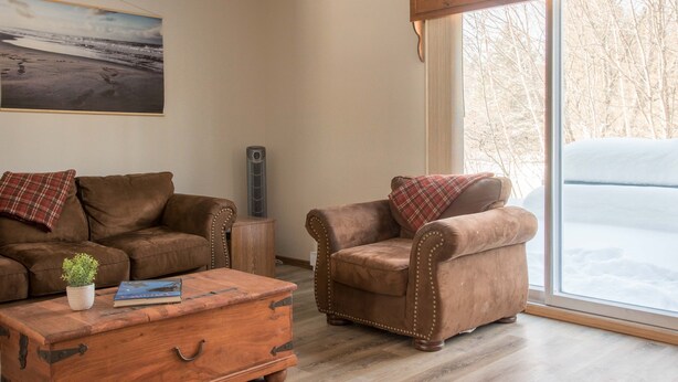 A cozy living room with a brown leather sofa, a matching armchair, and a wooden coffee table. A painting of a beach scene adorns the wall, and a large window offers a view of a snowy landscape.