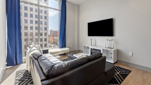 Ett modernt vardagsrum med en svart lädersoffa, ett vitt sidobord, en platt-TV som är monterad på väggen och ett stort fönster med blå gardiner.
