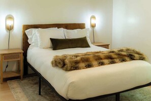 Una cama con colchón blanco, dos almohadas, un cubrefrío de piel sintética y dos mesitas de noche con lámparas de pared.