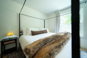 A bedroom with a four-poster bed, a bedside lamp, and a view of greenery outside.