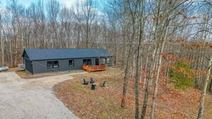 A two-story house with a dark gray exterior and a sloped roof, surrounded by a wooded area with fallen leaves.