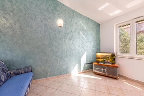 A room with a blue textured wall, a television on a wooden stand, a sofa with patterned cushions, and a window with a view of greenery.