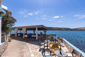 An outdoor dining area with stone flooring, tables, chairs, and a covered seating area overlooking a body of water.