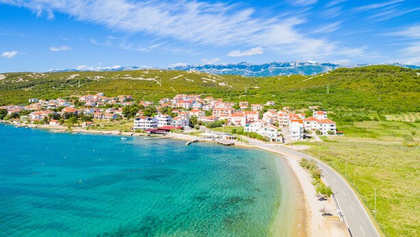 Une ville côtière bordée d’une mer d’un bleu éclatant, avec une route longeant le rivage et des bâtiments aux toits rouges.