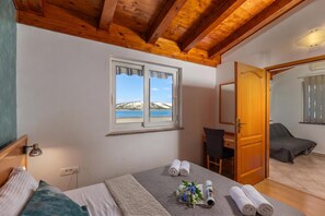 A bedroom with a wooden ceiling, a window with a view of a beach, a bed with pillows and a blanket, a chair, and a door leading to another room.