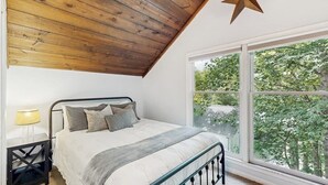 A bedroom with a wooden ceiling, a large window with a view of trees, a bed with pillows, and a nightstand with a lamp. - Charming dog-friendly cottage near Blowing Rock with private hot tub, deck, BBQ (Blowing Rock)