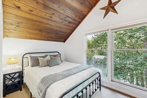 Une chambre avec un plafond en bois, une grande fenêtre donnant sur les arbres, un lit avec des oreillers et une table de chevet avec une lampe.