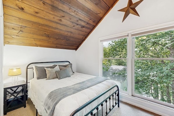 A bedroom with a wooden ceiling, a large window with a view of trees, a bed with pillows, and a nightstand with a lamp.