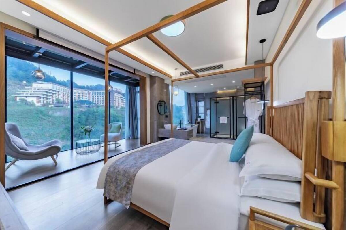 Una habitación de hotel moderna con una cama grande, un área de descanso con una silla y vistas a un edificio a través de amplios ventanales.