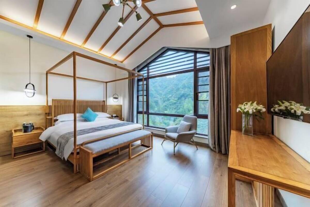 Un dormitorio moderno con una cama con dosel, un banco de madera, una silla, una mesita de noche y un amplio ventanal con vistas a zonas verdes.