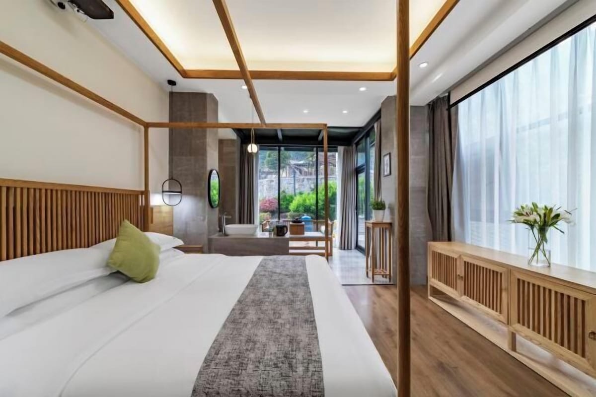 A modern bedroom with a large bed, a wooden headboard, a bedside table, a nightstand, a window with curtains, and a view of greenery outside.