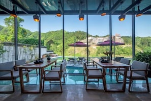 Un comedor con mesas y sillas, una piscina rodeada de cristal y vistas a un edificio en una colina.