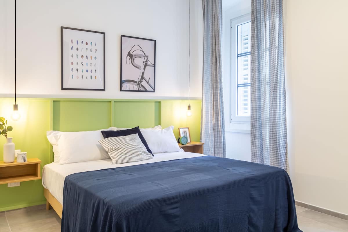 A neatly arranged bedroom with a green headboard, a large bed with a dark blue bedspread, two framed artworks on the wall, and a window with checkered curtains.
