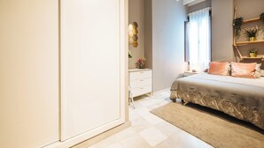 Een slaapkamer met een bed, nachtkastje, dressoir, raam met gordijnen en een wandplank met planten.