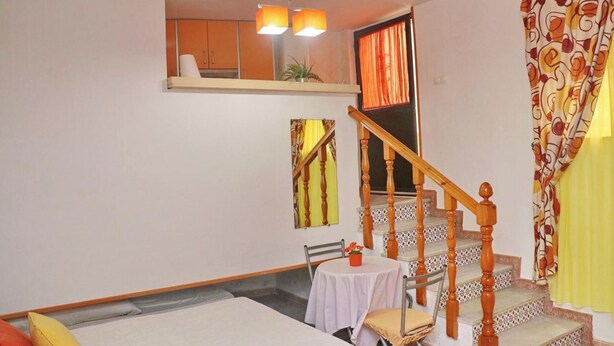 A room with a bed, a small table with a chair, a staircase with wooden railings, and a window with curtains.