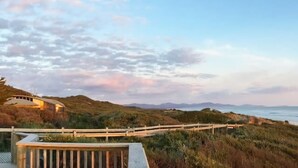 A coastal landscape with a wooden deck overlooking a grassy area and a building with a curved roof. - Sandy Point Log Cabin (Sandy Point)