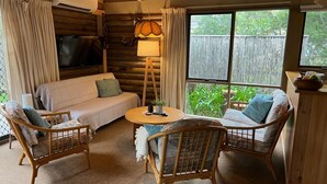 A cozy log cabin interior with a sofa, two wicker chairs, a small round table, and a window with a view of greenery. - Sandy Point Log Cabin (Sandy Point)