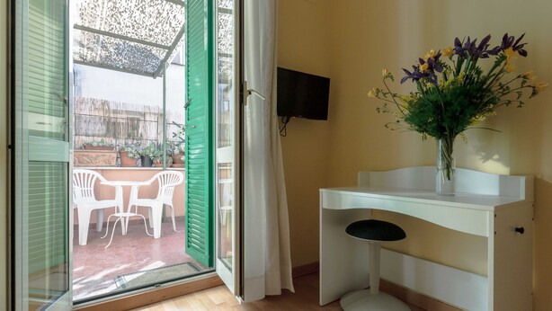 Eine offene Tür zu einem Außenbereich mit Tisch und Stühlen, einem weißen Schreibtisch mit Fernseher und einer Vase mit Blumen.