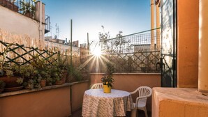 Eine kleine Terrasse im Freien mit einem runden Tisch und zwei Stühlen, umgeben von Topfpflanzen und einem Metallzaun.