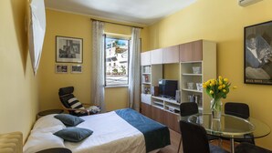 Ein gemütliches Hotelzimmer mit Bett, kleinem Schreibtisch, Stuhl, Glastisch und Blick auf die Straße durch ein Fenster.