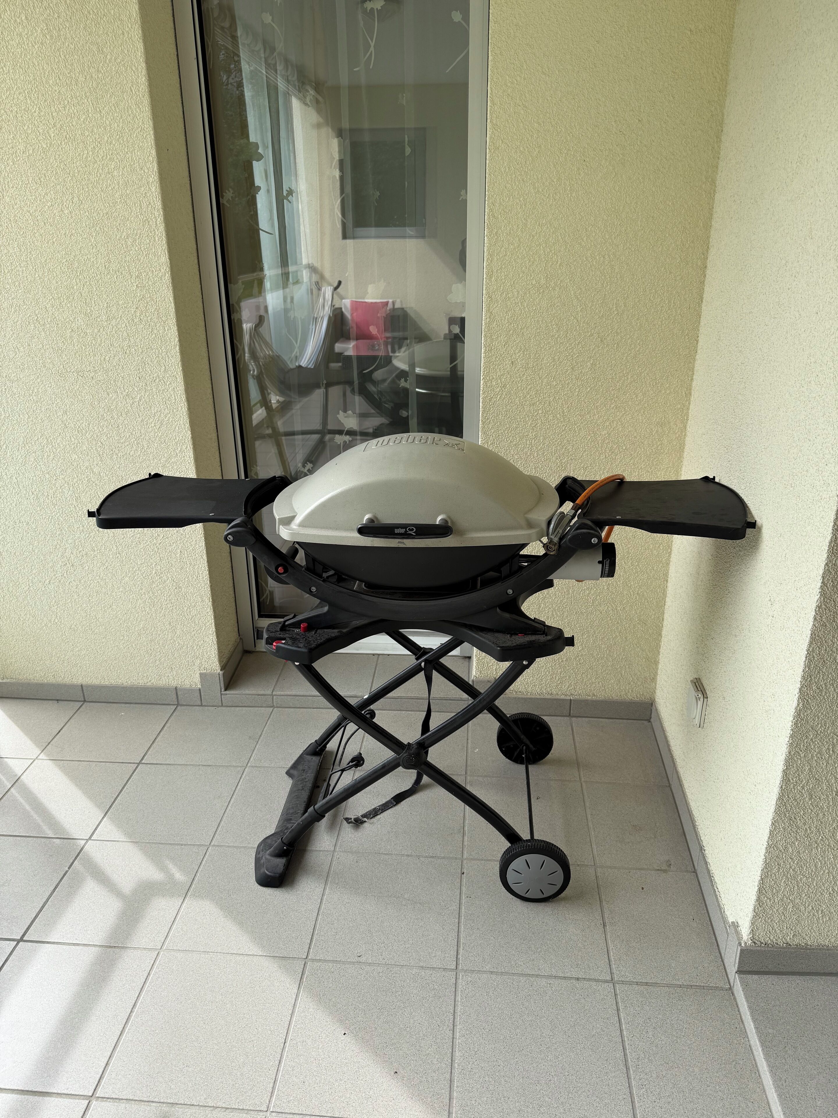 A portable grill with wheels, placed on a tiled floor near a glass door.