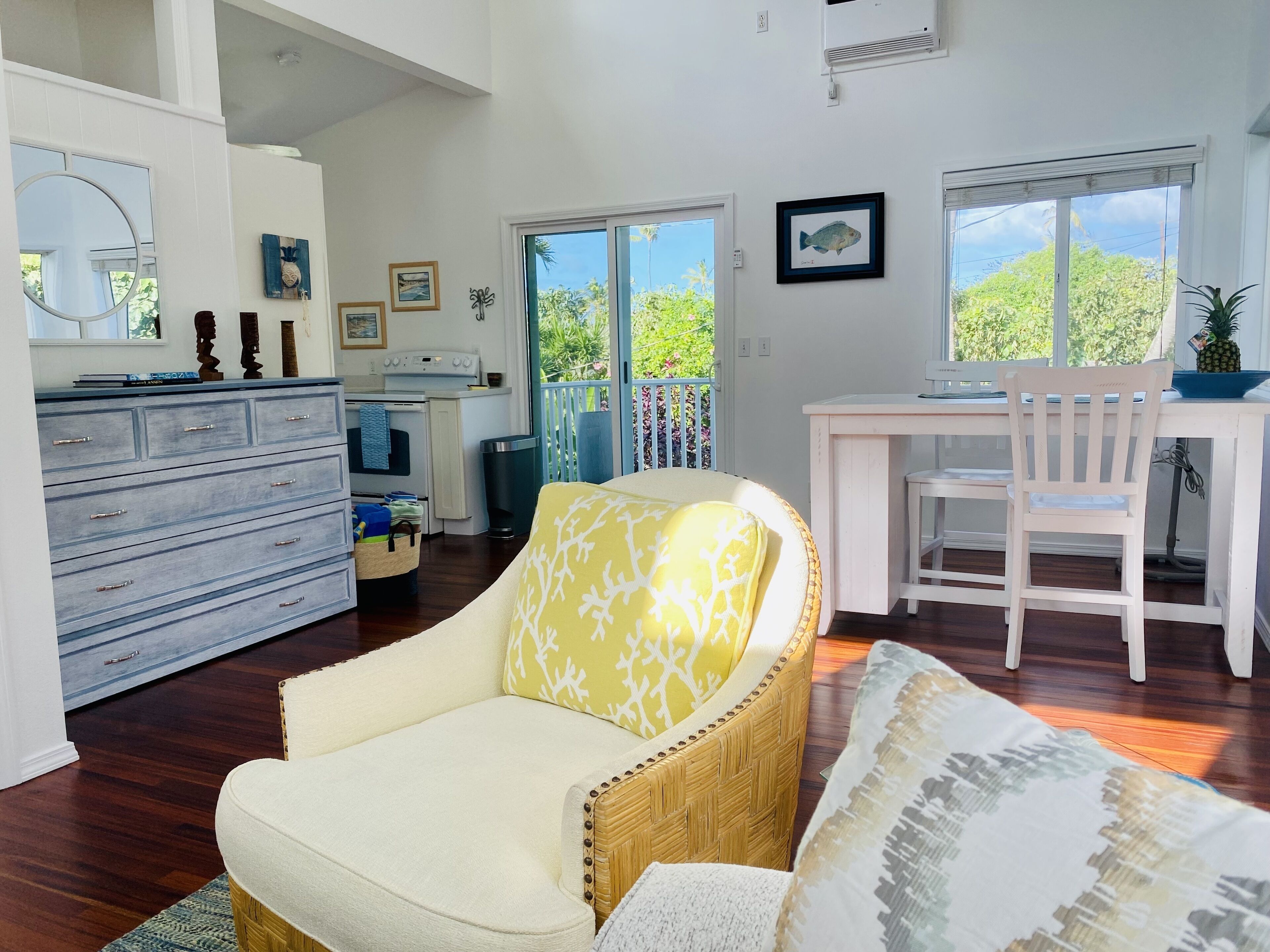 A spacious living area with a dining table, a wicker chair, and a sofa with a patterned cushion. A kitchen with a stove and a trash can is visible in the background.