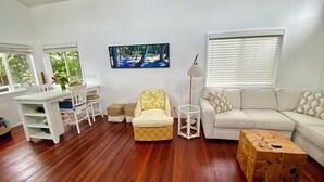 A living room with a white sofa, a white coffee table, a wooden side table, a white desk with a chair, and a painting on the wall.