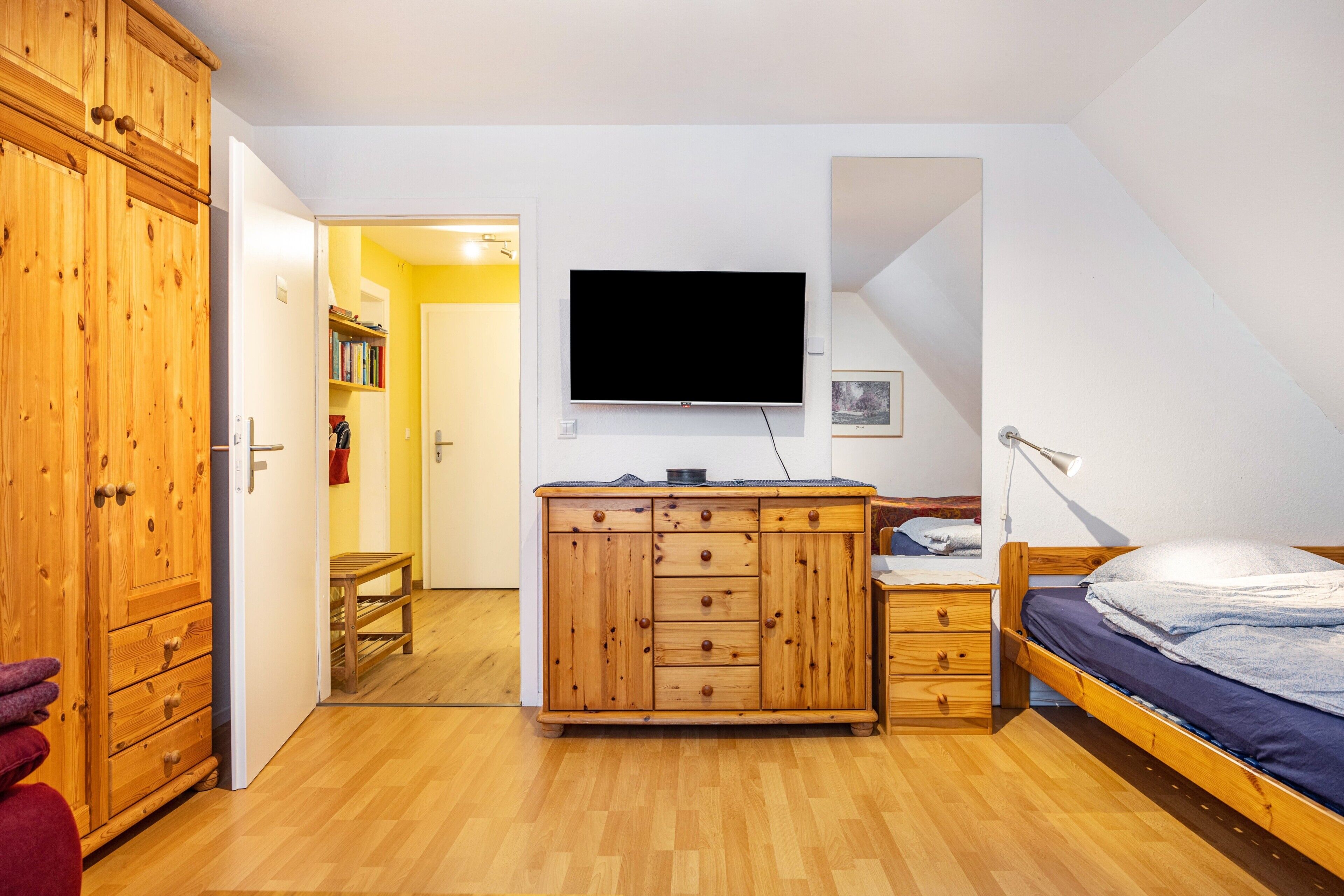 Ein Schlafzimmer mit Holzschrank, Bett, an der Wand montiertem Fernseher und kleinem Schreibtisch mit Stuhl.